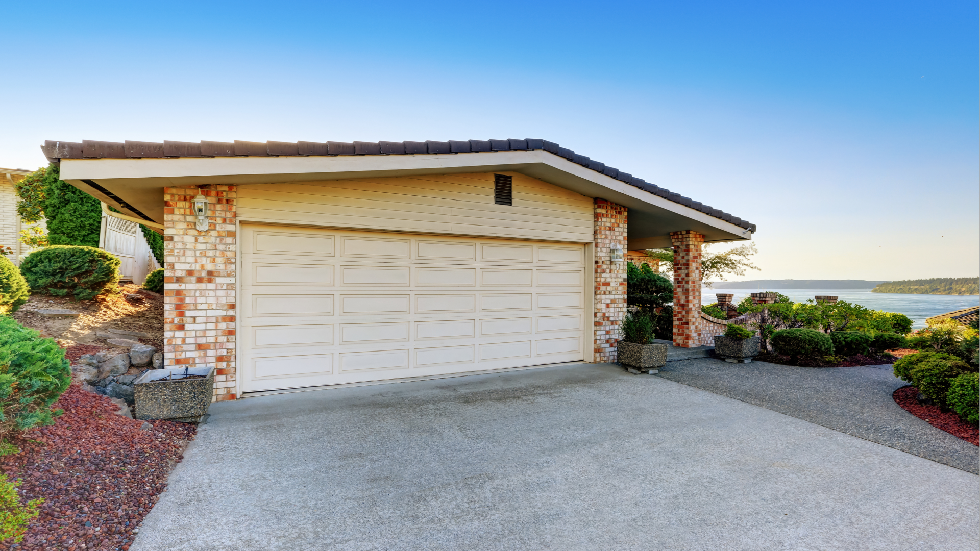Garage with white door, tan walls, and a stone facade, situated on a concrete driveway with waterfront views.
