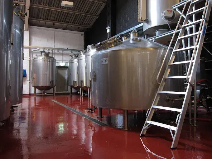 Brewery interior with stainless steel tanks and a red floor. A ladder leans against one of the tanks.
