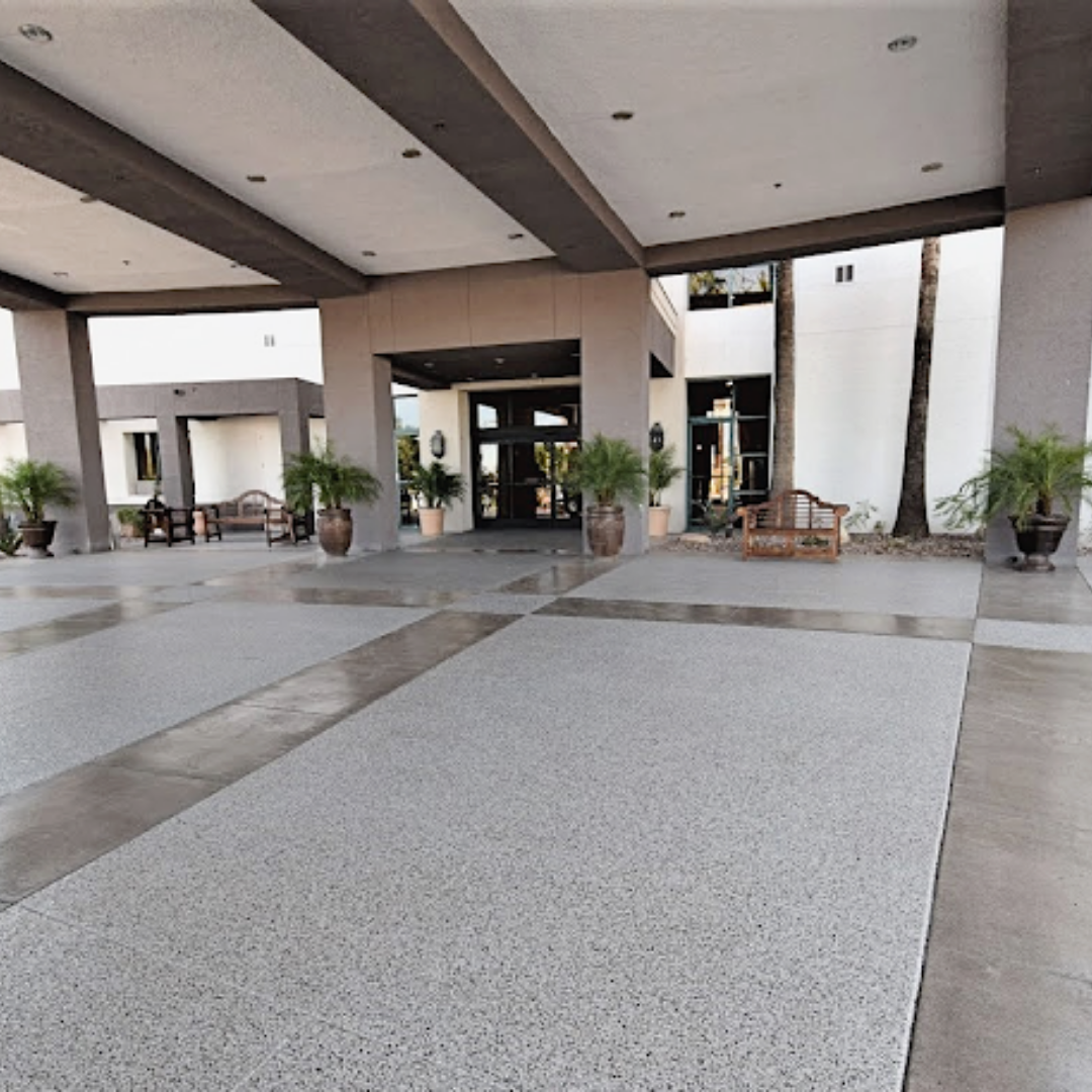Exterior of building with large covered entrance, terrazzo flooring, potted plants, and wooden benches.